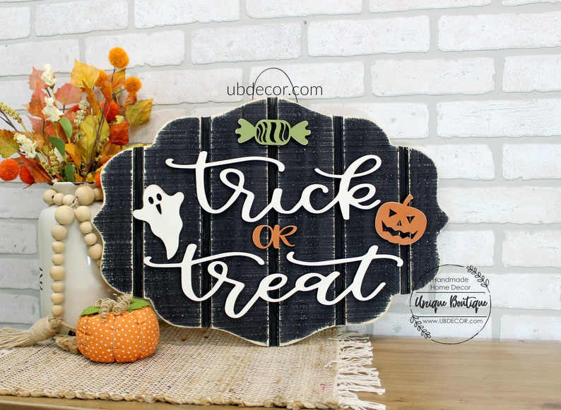 May include: Black wooden sign with the words "trick or treat" in white script. The sign is decorated with a white ghost, a green candy, and an orange pumpkin with a jack-o-lantern face. The sign is hanging on a white brick wall.