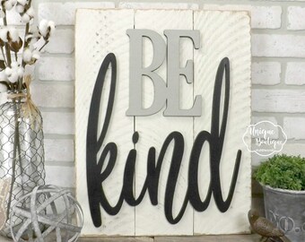 May include: White wooden sign with the words "BE KIND" in black and gray letters. The sign is distressed and has a rustic look.