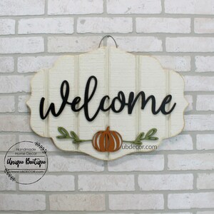 May include: Welcome sign with a white background and black lettering. The sign has a pumpkin and leaves in the center. The sign is hanging on a brick wall.