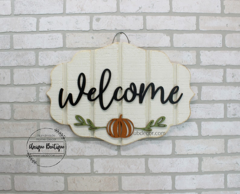May include: Welcome sign with a white background and black lettering. The sign has a pumpkin and leaves in the center. The sign is hanging on a brick wall.