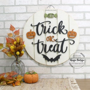 May include: A round wooden sign with the words "trick or treat" in black lettering. The sign is decorated with two orange pumpkins, a bat, and a green candy. The sign is hanging on a white brick wall.