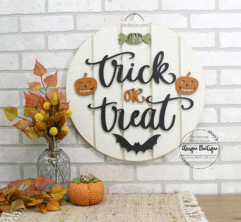 May include: A round wooden sign with the words "trick or treat" in black lettering. The sign is decorated with two orange pumpkins, a bat, and a green candy. The sign is hanging on a white brick wall.