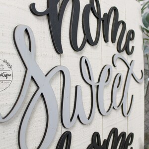 May include: A wooden sign with the words "Home Sweet Home" in black and gray layered lettering. The sign is on a whitewashed wood background.