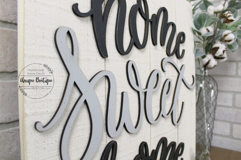 May include: A wooden sign with the words "Home Sweet Home" in black and gray layered lettering. The sign is on a whitewashed wood background.
