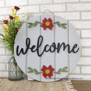 Pu&ograve; includere: Un cartello di benvenuto rotondo in legno con uno sfondo a strisce grigio e bianco. La parola "benvenuto" &egrave; scritta in corsivo nero. Il cartello &egrave; decorato con fiori rossi e gialli e foglie verdi.