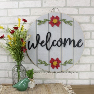 Pu&ograve; includere: Un cartello rotondo in legno con sfondo grigio e strisce bianche dipinte. Il cartello dice "welcome" in lettere di scrittura nere. Il cartello &egrave; decorato con fiori rossi e gialli e foglie verdi.