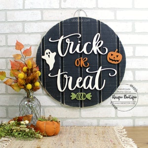 May include: A black and white wooden round sign with the words "trick or treat" in white script. The sign has a ghost, a pumpkin, and a candy cane on it. The sign is hanging on a white brick wall.