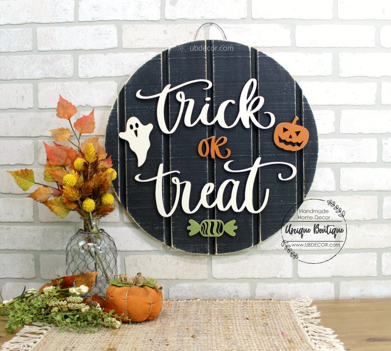 May include: A black and white wooden round sign with the words "trick or treat" in white script. The sign has a ghost, a pumpkin, and a candy cane on it. The sign is hanging on a white brick wall.