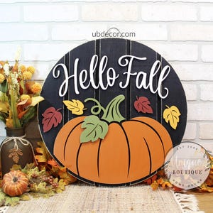 May include: A round black wooden sign with vertical slats featuring the white text Hello Fall. A large orange pumpkin with a green stem and leaves is centered below the text, accented by small red and yellow leaves on the sides.