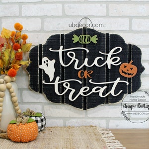 May include: Black wooden sign with white lettering that reads "trick or treat". The sign is decorated with a white ghost, a green candy, and an orange pumpkin. The sign is hanging on a white brick wall.