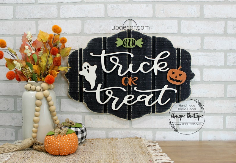 May include: Black wooden sign with white lettering that reads "trick or treat". The sign is decorated with a white ghost, a green candy, and an orange pumpkin. The sign is hanging on a white brick wall.