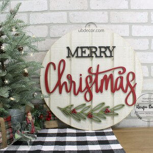 K&ouml;nnte beinhalten: Ein rundes Holzschild mit wei&szlig;em Hintergrund und den Worten "Merry Christmas" in roten und schwarzen Buchstaben. Das Schild ist mit einem gr&uuml;nen und roten Stechpalmenzweig verziert.