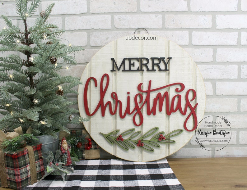 K&ouml;nnte beinhalten: Ein rundes Holzschild mit wei&szlig;em Hintergrund und den Worten "Merry Christmas" in roten und schwarzen Buchstaben. Das Schild ist mit einem gr&uuml;nen und roten Stechpalmenzweig verziert.