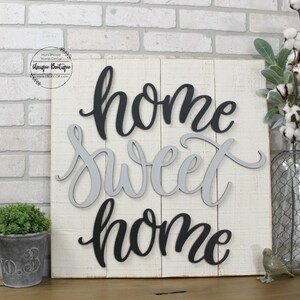 May include: A white wooden sign with the words "home sweet home" written in black and gray letters. The letters are layered and have a 3D effect.