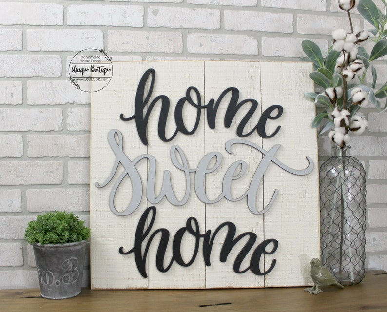 May include: A white wooden sign with the words "home sweet home" written in black and gray letters. The letters are layered and have a 3D effect.