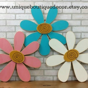 Puede incluir: Tres decoraciones de pared de madera en forma de margarita en rosa, turquesa y blanco con un dise&ntilde;o de espiral dorado en el centro de cada flor.