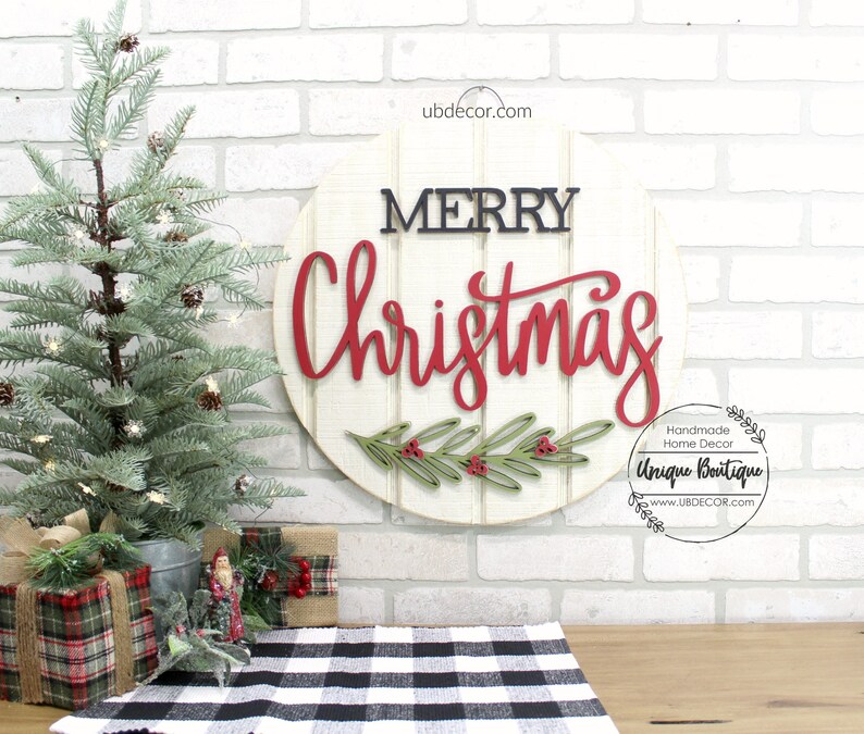 K&ouml;nnte beinhalten: Ein rundes Holzschild mit den Worten "Merry Christmas" in roten und schwarzen Buchstaben. Das Schild ist mit einem gr&uuml;nen Blattzweig mit roten Beeren verziert. Das Schild befindet sich vor einem wei&szlig;en Backsteinmauer Hintergrund.