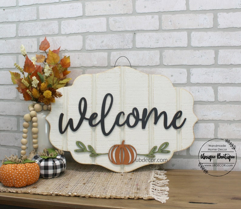 May include: A white wooden welcome sign with black cursive lettering and a cutout orange pumpkin with green leaves. The sign has a distressed look and is hanging from a metal hook. The sign is on a table with a brown wooden surface and a beige rug.