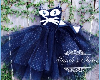 May include: Black tulle tutu with white polka dots and a black cat costume top with white ears and a white bow. The costume includes a black headband with white cat ears.