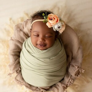 May include: A newborn baby girl is sleeping peacefully in a light brown basket. She is wrapped in a light green swaddle and wearing a headband with peach and white flowers.