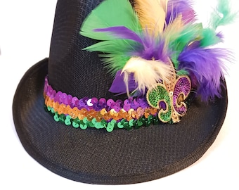 May include: A black fedora hat with a green, purple and gold sequin band. The hat is decorated with a gold fleur-de-lis pin and a cluster of green, purple and white feathers.