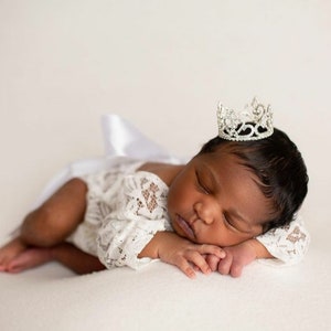 May include: A newborn baby wearing a white lace outfit and a silver tiara, sleeping on a white surface.