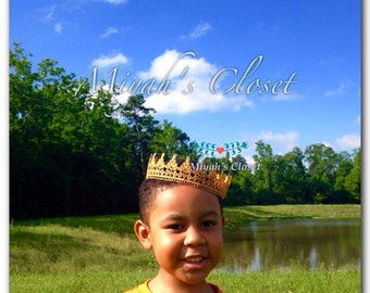 May include: A young boy wearing a gold crown, smiling at the camera. The crown is made of crocheted yarn and has a pointed top. The boy is wearing a bright orange shirt and is standing in a grassy field with a pond in the background. The text "Miyah's Closet" is visible in the image.