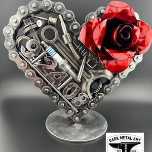 Puede incluir: Una escultura de metal en forma de corazón hecha de una cadena, herramientas y una rosa roja. El corazón está de pie sobre una base de metal. La escultura está firmada "Dark Metal Art" con el texto "EST 2013" y "HAND MADE".