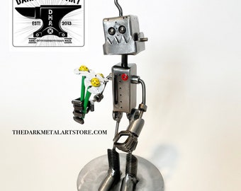 May include: A silver robot holding a bouquet of white flowers with yellow centres. The robot is made of metal and has a red button on its chest. The robot is standing on a round metal base. The robot is holding the flowers in its left hand and its right hand is raised in a gesture of greeting. The robot is a handmade metal sculpture.