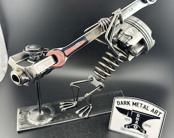 Puede incluir: Escultura metálica de una figura similar a un pájaro, hecha con piezas de automóviles. La escultura presenta una llave, un pistón y un resorte, con una base rectangular. Una pegatina con el texto "DARK METAL ART" es visible.