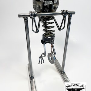 May include: A metal sculpture of a skeleton doing a pull-up on a bar. The skeleton is made of recycled metal parts, including a piston for the head. The sculpture is mounted on a metal stand. The sculpture is signed "DARK METAL ART ESTI 2013 HAND MADE"
