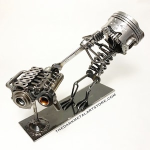 May include: A metal sculpture of a skeleton lifting a car engine with a wrench. The sculpture is made of recycled metal parts and is mounted on a black base. The base has the text "THEDARKMETALARTSTORE.COM" on it.