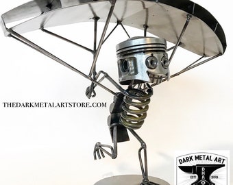 May include: A unique metal sculpture of a figure paragliding. The figure's head is a piston, and its body is made of metal rods and springs. The paraglider is made of metal sheets. The sculpture is on a round metal base. TheDarkMetalArtStore.com is written on the left.