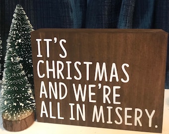 May include: A brown wooden sign with white text that reads "It's Christmas and we're all in misery."