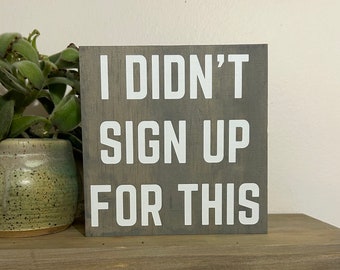 May include: A gray wooden sign with white lettering that reads "I DIDN'T SIGN UP FOR THIS." The sign is displayed on a wooden shelf, with a green plant in a ceramic pot to the left.