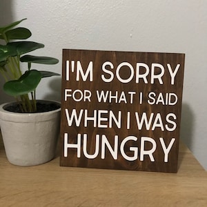 May include: A wooden sign with the text "I'M SORRY FOR WHAT I SAID WHEN I WAS HUNGRY" in white lettering. The sign is dark brown and sits on a wooden surface. A small potted plant is to the left of the sign.