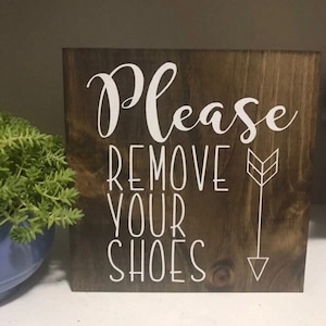 May include: A wooden sign with the text "Please Remove Your Shoes" in white lettering. The sign has a brown wood grain and a white arrow pointing down.