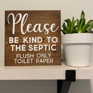 May include: A wooden sign with white text that reads "Please be kind to the septic flush only toilet paper". The sign is sitting on a white shelf with a small green plant in a white pot to the right.