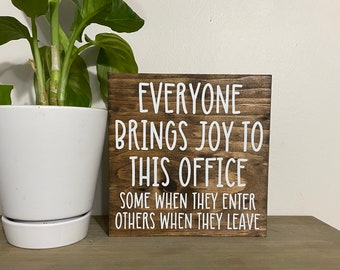 May include: A brown wooden sign with white text that reads "Everyone brings joy to this office. Some when they enter, others when they leave."