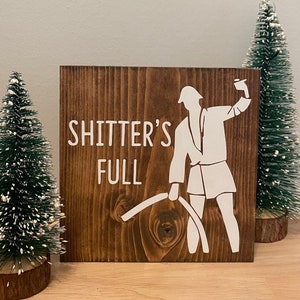 May include: A brown wooden sign with white text that reads "Shitter's Full". A white silhouette of a person holding a roll of toilet paper is on the sign.
