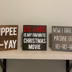 May include: Three wooden signs with white text on a shelf. The signs read: "YIPPEE KI-YAY", "DIE HARD IS MY FAVORITE CHRISTMAS MOVIE", and "NOW I HAVE A MACHINE GUN HO-HO-HO". The signs are brown, black, and gray.