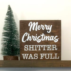 May include: A small, dark brown wooden sign with white lettering that reads "Merry Christmas SHITTER WAS FULL." A small green Christmas tree sits to the left of the sign. The sign is on a brown shelf.