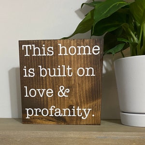 May include: A wooden sign with the text "This home is built on love & profanity." The sign is brown with white lettering and has a rustic look.