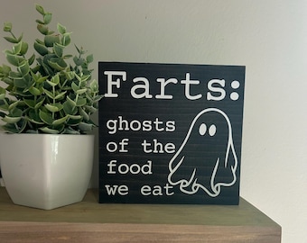 May include: A black wooden sign with white text that reads "Farts: ghosts of the food we eat" and a white ghost illustration. A small green plant in a white pot is to the left of the sign. The sign is on a wooden shelf.