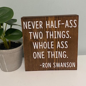 May include: A wooden sign with the quote "Never half-ass two things. Whole ass one thing. -Ron Swanson" in white lettering.