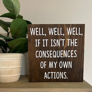 May include: A dark brown wooden sign with white text that reads "WELL, WELL, WELL, IF IT ISN'T THE CONSEQUENCES OF MY OWN ACTIONS."