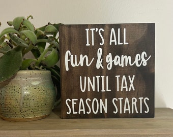 May include: A brown wooden sign with white text that reads "It's all fun & games until tax season starts".
