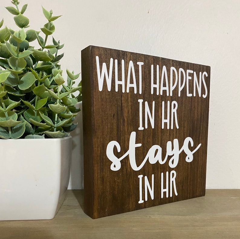 May include: A brown wooden sign with white text that reads "What happens in HR stays in HR".