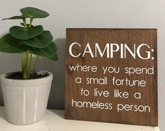 May include: A brown wooden sign with white text that reads "Camping: where you spend a small fortune to live like a homeless person."
