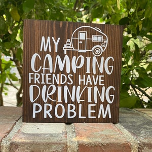 May include: A brown wooden sign with white text that reads "My camping friends have a drinking problem". A white silhouette of a camper is above the text.